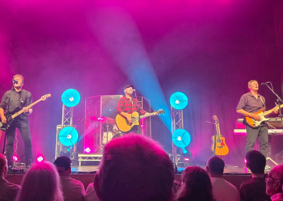 A live band performs on stage under vibrant pink and blue lighting, with four musicians visible playing guitars, bass, and percussion. Behind them are also a drummer and a keyboard player