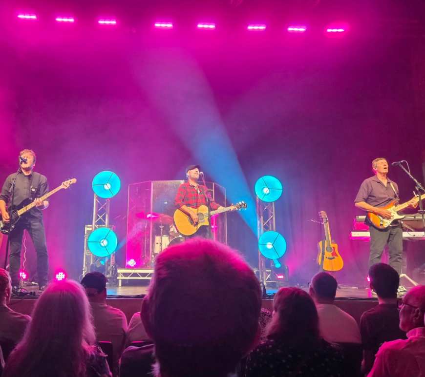 A live band performs on stage under vibrant pink and blue lighting, with four musicians visible playing guitars, bass, and percussion. Behind them are also a drummer and a keyboard player